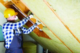 L'image représente un homme, un ouvrier, qui installe une mousse dans un mur. La scène montre donc des travaux pour une maison individuelle par un ouvrier qui porte un casque et un masque de chantier ainsi qu'une tenue de chantier.