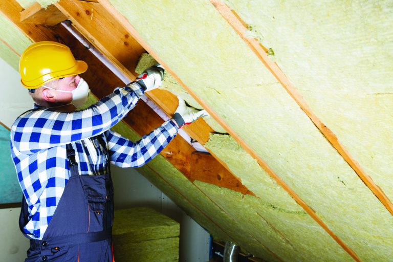 L'image représente un homme, un ouvrier, qui installe une mousse dans un mur. La scène montre donc des travaux pour une maison individuelle par un ouvrier qui porte un casque et un masque de chantier ainsi qu'une tenue de chantier.